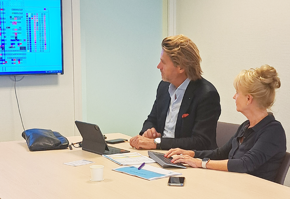 Twee personen zitten aan een vergadertafel en kijken naar een scherm met een digitaal rooster; op tafel liggen documenten, een tablet, een laptop en een telefoon.