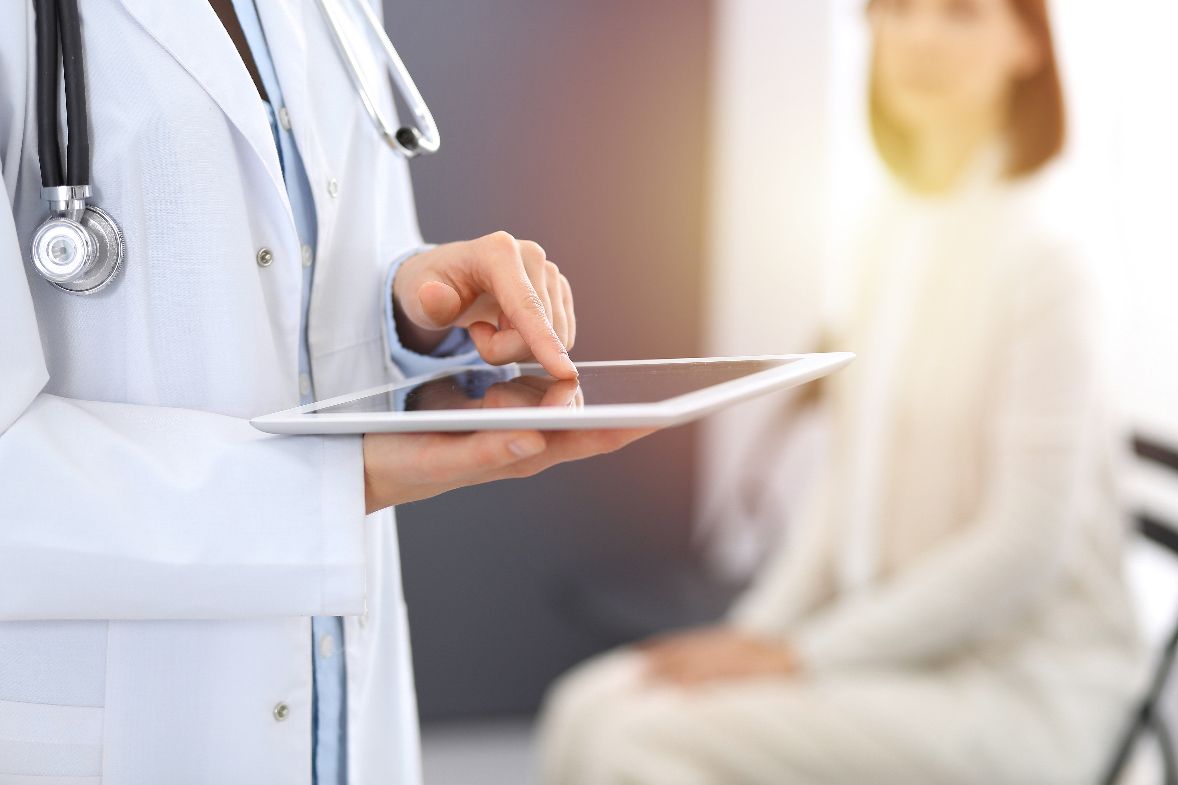 Een zorgverlener zit met zijn linkerhand op een tablet. Op de achtegrond zit een patiënt te wachten