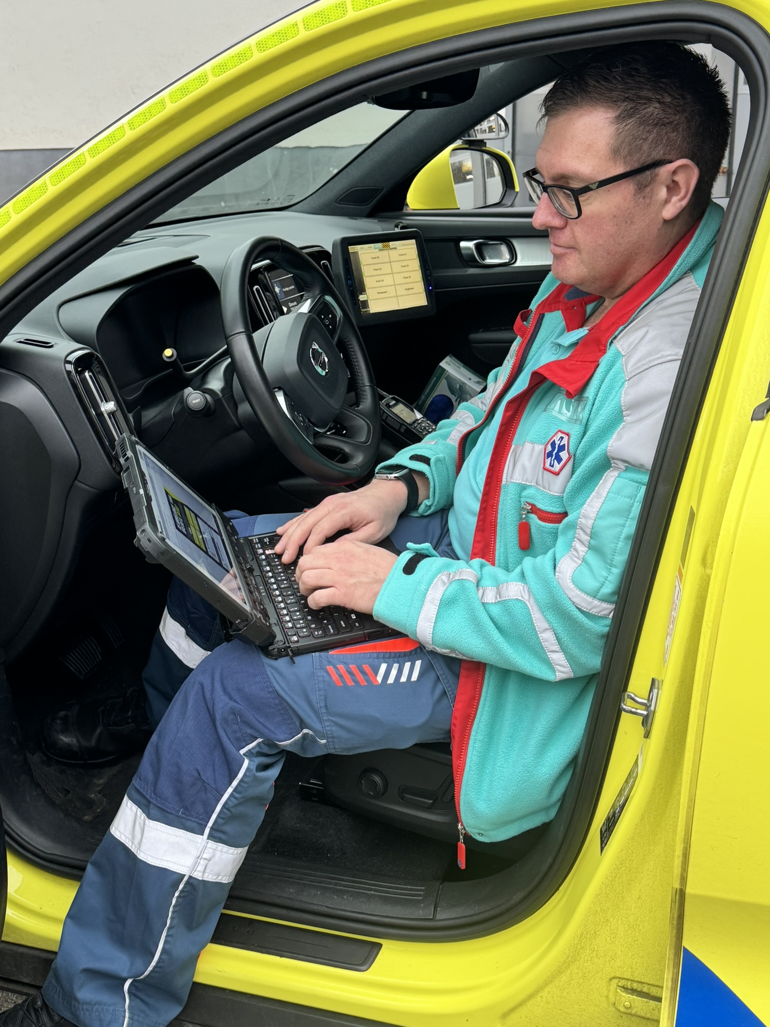 A healthcare professional is sitting in the driver’s seat of a yellow ambulance, working on a laptop placed on their lap, with the dashboard and an integrated screen visible in the background.
