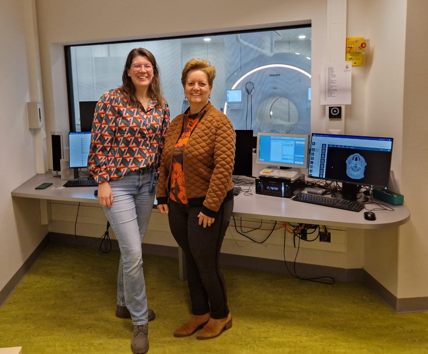 Zwei Personen stehen in einem Kontrollraum neben einem MRT‑Scanner, mit mehreren Bildschirmen und medizinischer Software im Hintergrund.