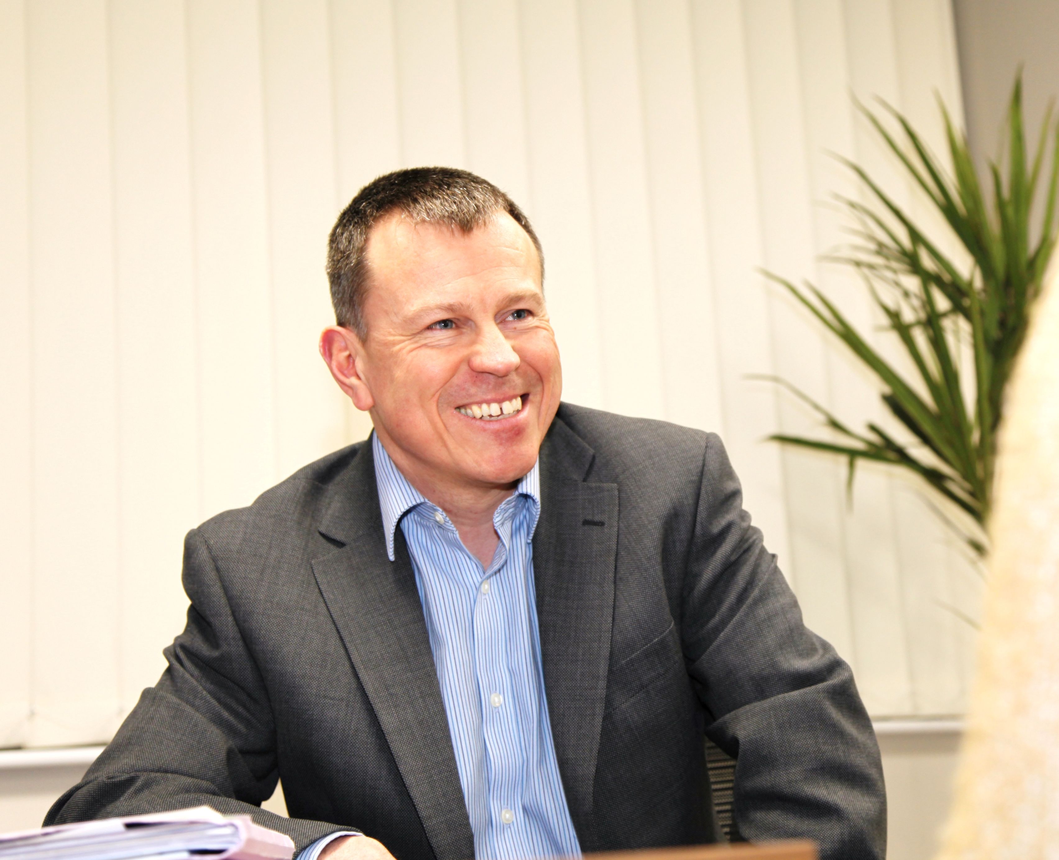A person seated at a desk in an office setting, wearing a suit jacket and a light blue shirt, with papers on the table and a plant in the background.