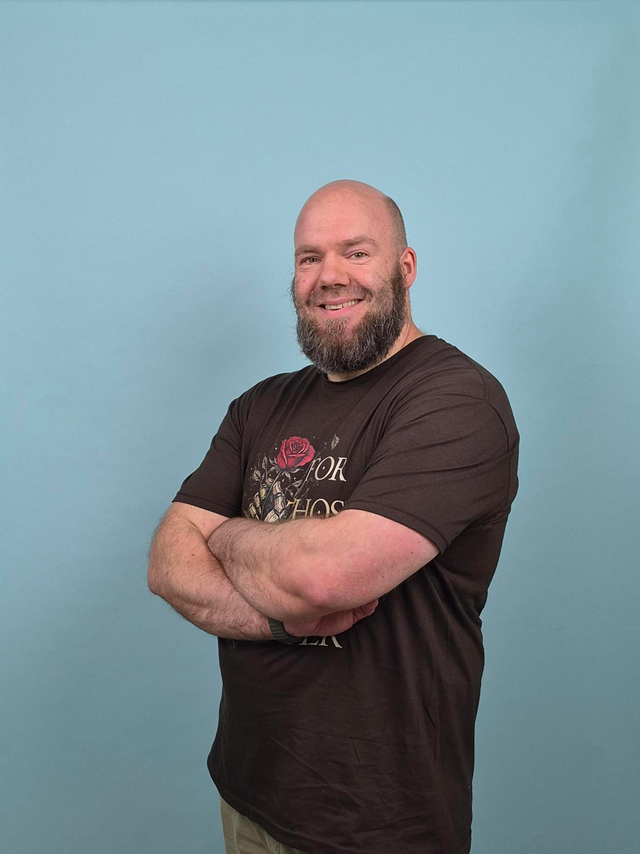 Person with arms crossed wearing a dark T-shirt featuring a rose graphic and partially visible text, standing against a plain light blue background.